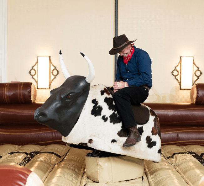 Man riding a mechanical bull at an indoor event, wearing a cowboy hat and red bandana, showcasing entertainment options for parties in Charleston.